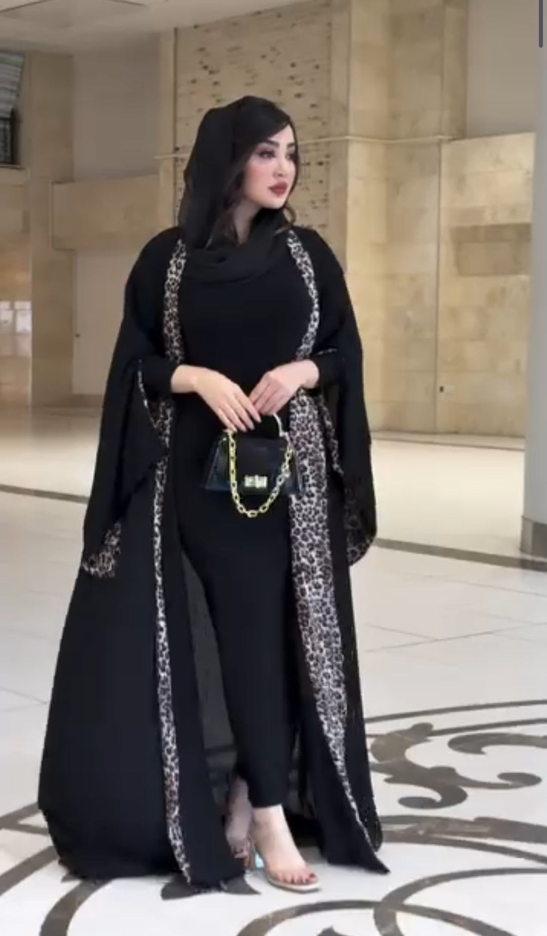 Woman in a black outfit with intricate designs standing in a modern indoor setting.