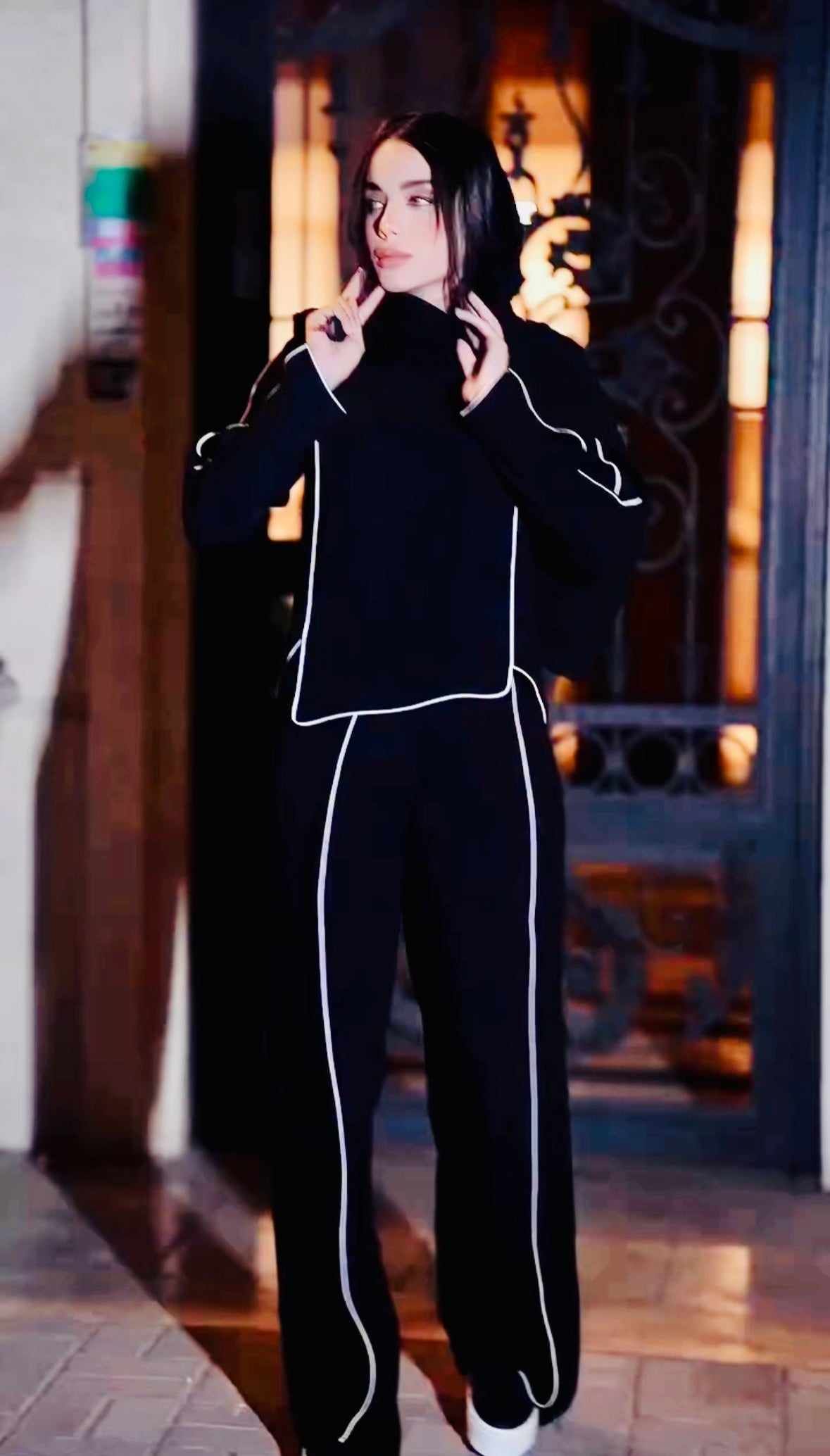 Person wearing a black outfit with white piping in a dimly lit room.